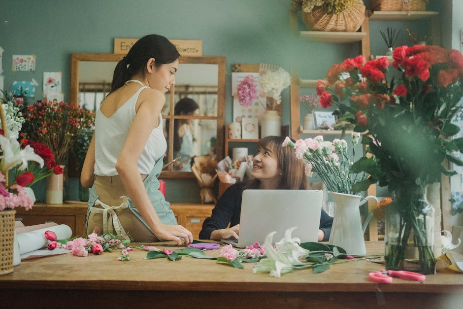 A female florist and a customer in a flower shop engaged in a conversation about floral arrangements. The florist, wearing a white sleeveless top and an apron, stands at a wooden counter with various 