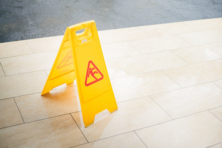 A bright yellow warning sign with red triangular symbols and a person slipping icon placed on a beige tiled floor, indicating a wet or slippery surface. The sign stands upright, with the tiles showing