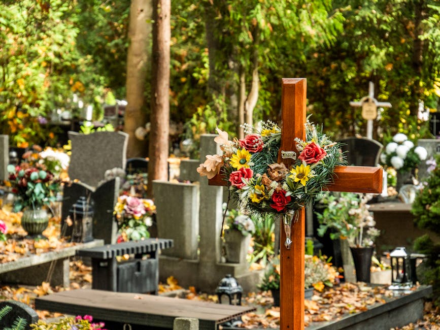 A wooden cross decorated with a floral arrangement resting at a gravesite in a cemetery. The arrangement includes red roses, yellow daffodils, pale pink lilies, and greenery, all fresh and vibrant. Su