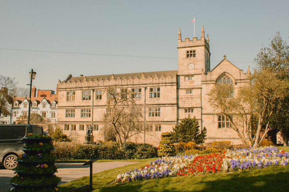 A vibrant floral display in front of a historic stone building featuring a tower and ornate architectural details, with a well-maintained garden bed full of colorful flowers including purple, red, and