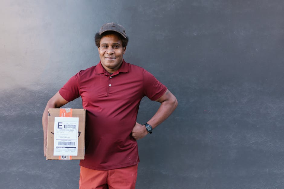 A smiling delivery person standing against a plain grey wall, holding a small cardboard parcel with an orange and white shipping label, dressed in a maroon polo shirt, red trousers, and a grey cap. Th