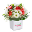 A spirit of lighthearted joy infuses this bouquet, displayed in a pristine white gift bag reminiscent of thoughtful surprises left on a neighbour's doorstep in West Byfleet. Coral and peach-toned carnations, each with delicately layered, frilly petals, exude warmth and affection-a familiar sight during springtime walks through Broadoaks Park. Crisp white daisies with cheerful, lime-fresh centers stand lively among the carnations, suggesting new beginnings and uplifting moods, while wisps of tiny white baby's breath wind gracefully amidst the blooms, adding softness and innocence. Along one side, hints of purple-tinged buds peek out, drawing the eye and offering the promise of further blossoms yet to open. The entire arrangement is hugged by verdant, glossy leaves, which fan out in elegant arcs to frame the bouquet, providing both contrast and structure. The ensemble rises upright from a sturdy white bag fitted with ribbon handles, its simplicity ensuring all focus remains on the floral artistry within. Illuminated by gentle, natural light, each vivid colour and lush texture seems to radiate with the happiness of shared moments-perfect for brightening a friend's day near West Byfleet village, marking a birthday, or celebrating a heartfelt milestone. Every stem is expertly placed, reflecting the genuine care and communal warmth that define floral gifts crafted in this corner of Surrey.